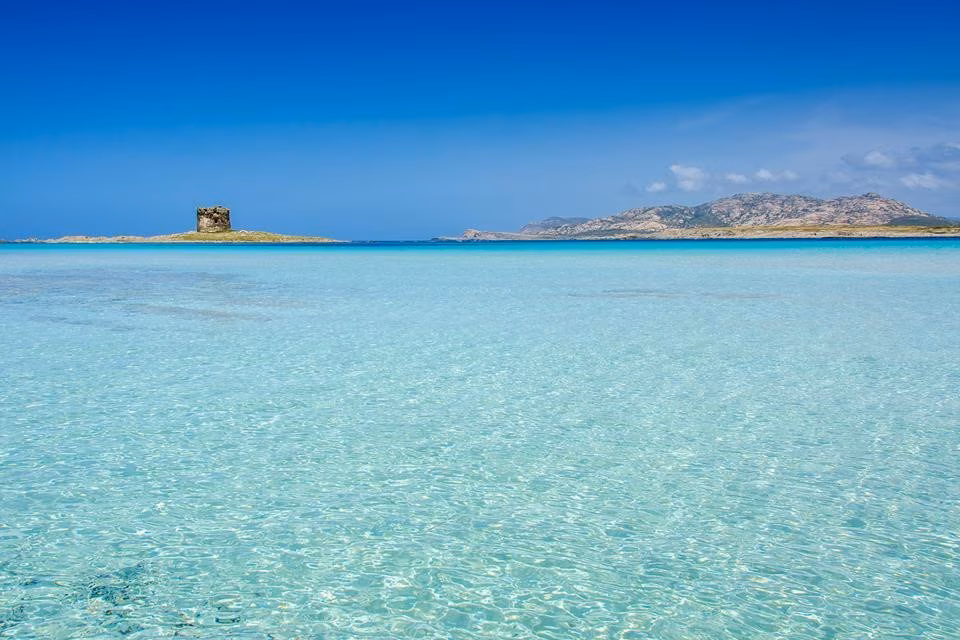 Stunning panoramic view of La Pelosa beach in Stintino, Sardinia, featuring shallow turquoise water, white sand, and the iconic Aragonese stone tower in the distance.