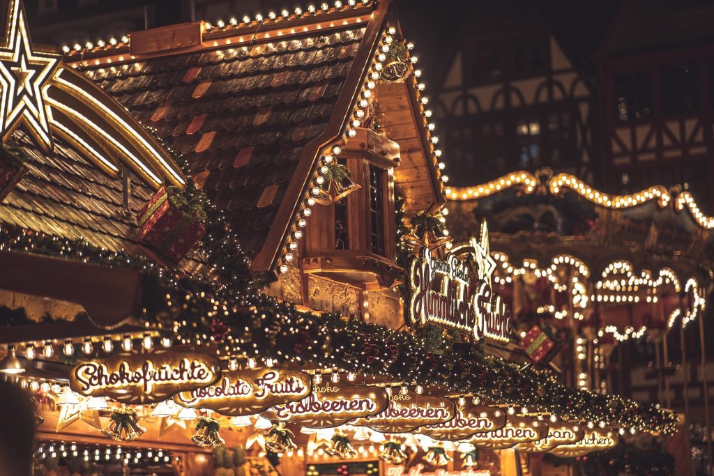 Christmas market stalls glowing with festive lights, warm decorations, cozy wooden rooftops, and a magical evening holiday atmosphere.