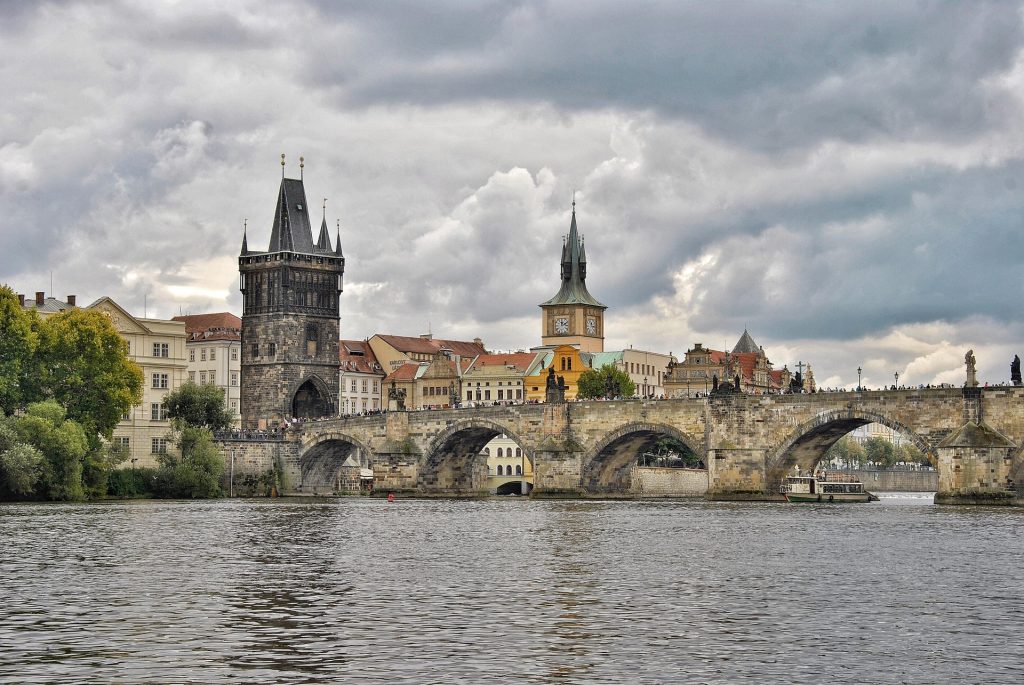 Historic stone bridge over the river with Gothic towers, dramatic clouds, and classic city architecture in the background.