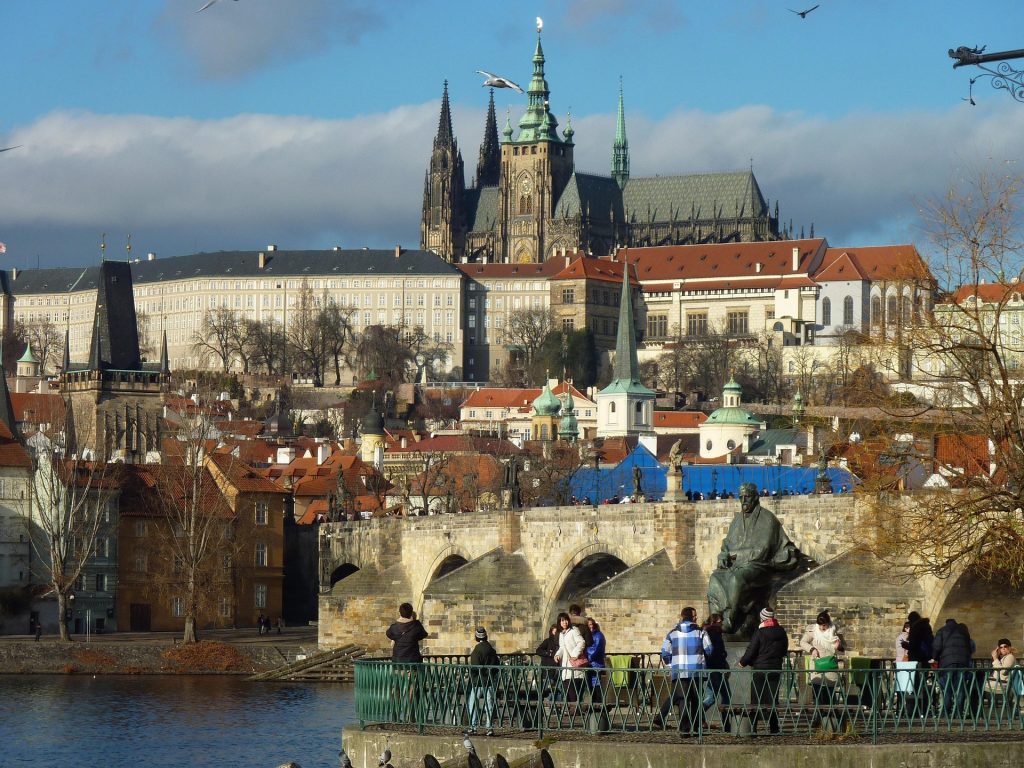 Hilltop castle complex with cathedral spires, red rooftops, riverfront promenade, and panoramic city views.