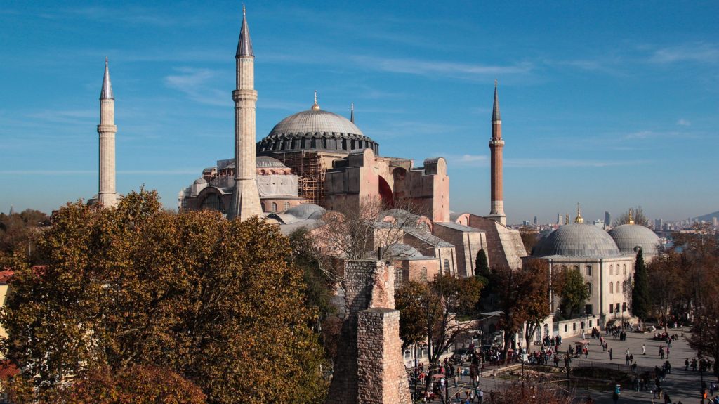 Daytime view of Hagia Sophia surrounded by historic domes, autumn trees, and the skyline of Istanbul in the background.