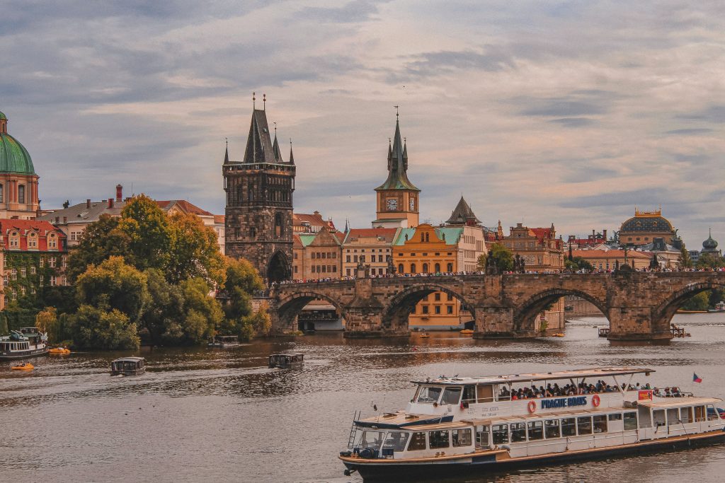 Historic stone bridge crossing the river with Gothic towers, elegant old buildings, and sightseeing boats passing through the city.