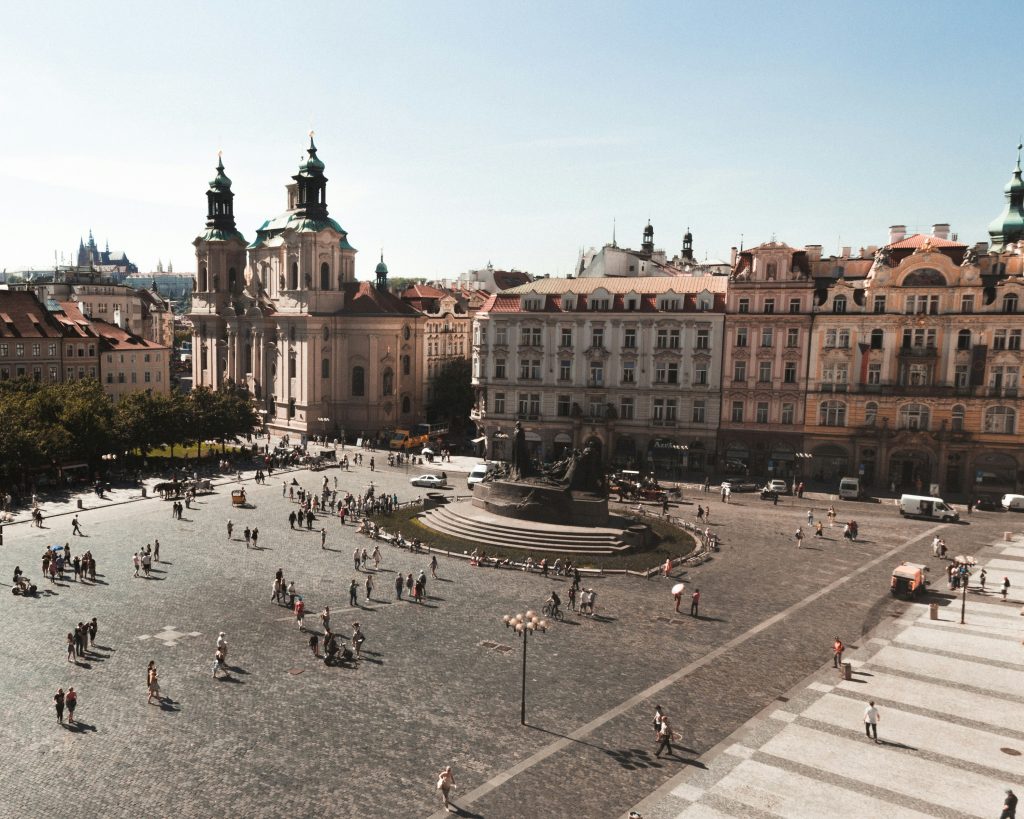 Wide historic square with colorful buildings, church towers, open cobblestone streets, and people walking through the city center.