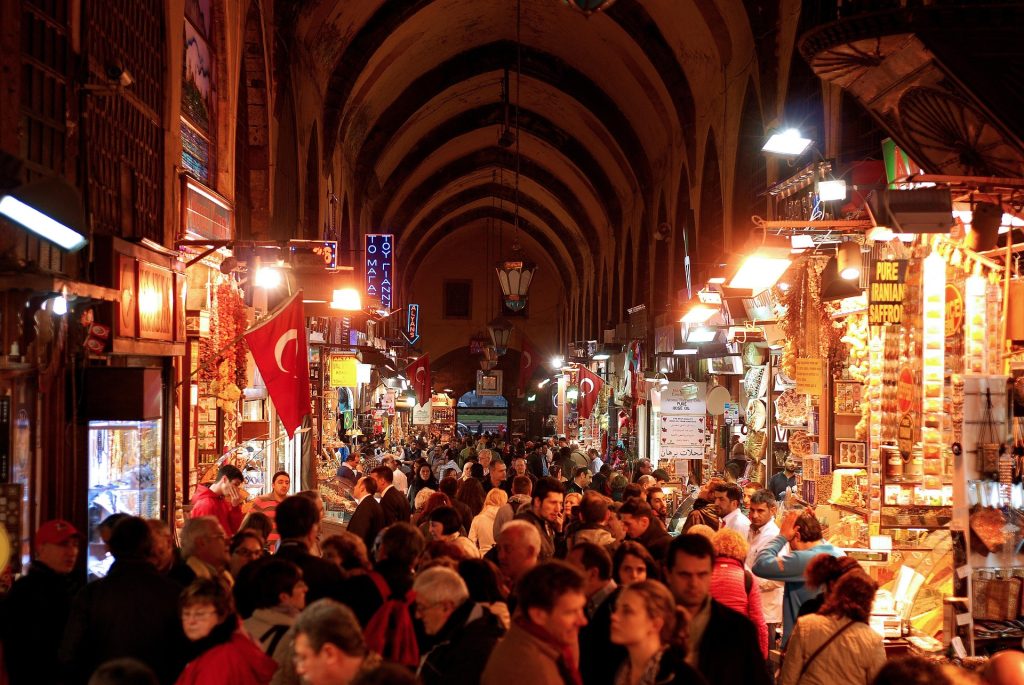 Crowded interior walkway of the Grand Bazaar filled with local shops, Turkish flags, warm lights, and busy evening shoppers.