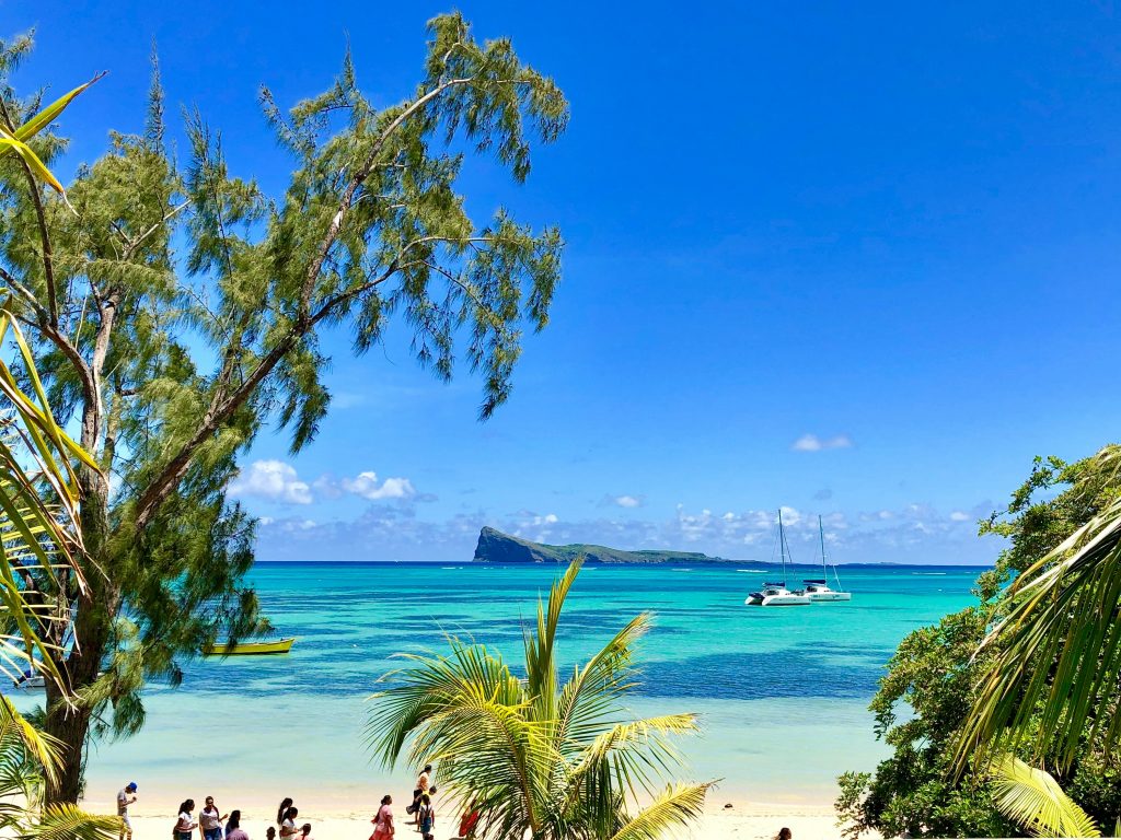 Palm trees framing a tropical beach with turquoise water, anchored sailboats, and a small island visible on the horizon